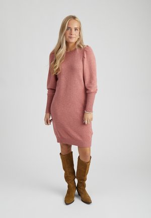 Mujer con cabello largo y rubio que lleva un vestido de punto rosa con mangas abullonadas y botas de ante marrones hasta la rodilla sobre un fondo liso.