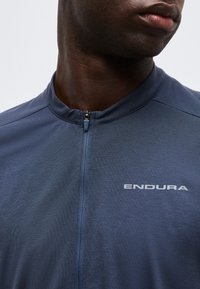 Κοντινό πλάνο ενός ατόμου που φοράει μπλε αθλητική μπλούζα με φερμουάρ και το λογότυπο «ENDURA» στο στήθος, με το φερμουάρ μερικώς κλειστό στον γιακά.