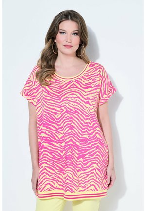Femme aux cheveux ondulés portant de grandes boucles d'oreilles hoop et un haut ample à manches courtes à rayures zébrées rose et jaune sur fond blanc.