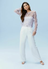 Transparente lila Bluse mit langen Ärmeln und floralen Applikationen, kombiniert mit hochgezogenen weißen weit geschnittenen Hosen und silbernen Sandalen mit offenem Zeh.