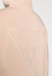 Ανοιχτό ροζ φούτερ με κουκούλα, με μεγάλο ανάγλυφο τριγωνικό λογότυπο στην πλάτη, π ου φέρει το κείμενο "GUESS U.S.A. WASHED JEANS" και λεπτομέρεια με ερωτηματικό.