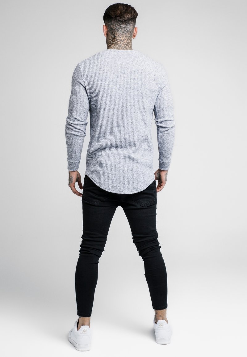 Hellgraues, geripptes Langarmshirt mit abgerundetem Saum, kombiniert mit schwarzen engen Jeans und weißen Sneakern, von hinten gesehen.