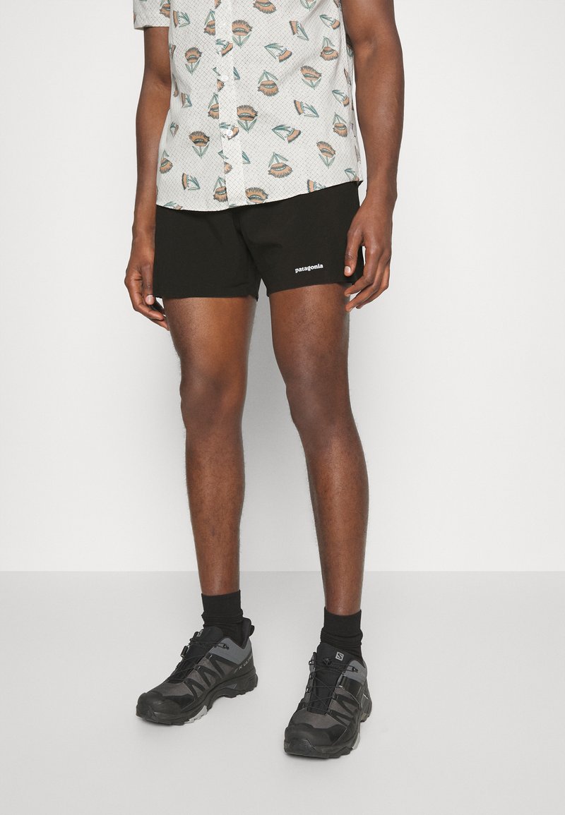Black Patagonia shorts with a printed white logo, worn with a patterned short-sleeve shirt and gray athletic shoes with black accents.
