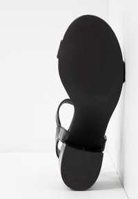 Semelle de sandale noire avec petit talon bloc et lanières texturées, présentée debout contre un coin blanc en arrière-plan.