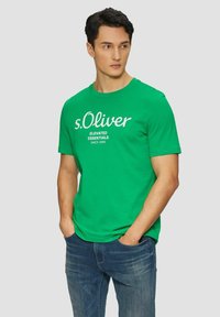 Grünes Baumwoll-T-Shirt mit Rundhalsausschnitt und kurzen Ärmeln, bedruckt mit "s.Oliver ELEVATED ESSENTIALS SINCE 1969" in weißer Schrift.