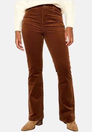 Femme portant un pantalon en velours côtelé marron et des bottines à talons brodées couleur beige, debout les mains détendues le long du corps.
