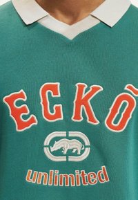Polo verde cu guler alb, având brodat în text portocaliu "ECKŌ" și "unlimited", precum și un grafic cu un rinocer în alb.