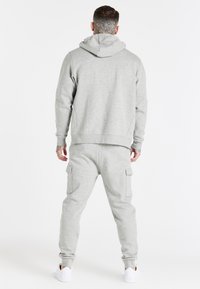 Felpa grigia con cappuccio e pantaloni coordinati in tessuto morbido, dotati di tasca frontale e tasche laterali in stile cargo, abbinati a scarpe da ginnastica bianche.