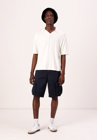 Camisa polo branca de mangas curtas e canelada, combinada com calções cargo azul-escuro com bolsos laterais; modelo usa um chapéu preto e ténis brancos.