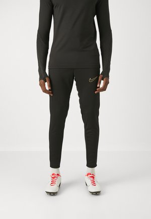Persoon die zwarte sportbroek en top draagt met een gouden Nike-logo, witte voetbalschoenen met rode veters, staand tegen een effen achtergrond.