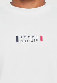 Valge sweatshirt tekstiilist, millel on ristkülikukujuline logo "TOMMY HILFIGER" mustade ja punaste aktsentidega rinnaosas.