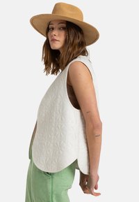 Chaleco blanco acolchado con un patrón floral, con dobladillo redondeado y sin mangas, llevado sobre una camiseta marrón y pantalones verdes. Combinado con un sombrero de paja de ala ancha.