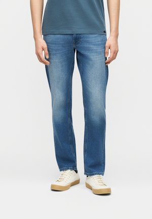 Bărbat purtând blugi albaștri, cu picior drept, decolorați, adidași albi cu talpă de cauciuc și un tricou albastru, stând în picioare pe un fundal simplu.