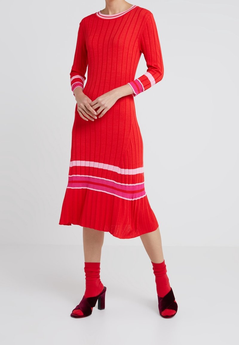 Robe rouge côtelée à manches longues avec des accents rayés rose et blanc, ourlet évasé. Associée à des sandales à talons bordeaux et des chaussettes rouges.