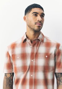 Camicia a maniche corte a quadri arancione e bianca con colletto abbottonato, due tasche sul petto e tessuto morbido e strutturato.