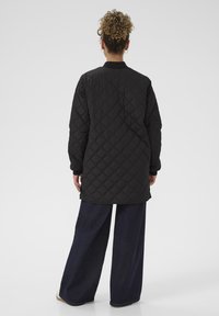Giacca lunga nera trapuntata con colletto arrotondato, vestibilità comoda e maniche lunghe, abbinata a pantaloni in denim scuro a gamba larga.