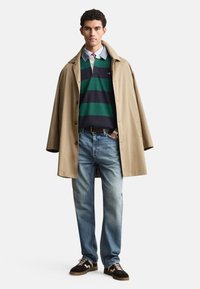 Bēšs mētelis virs svītrainas zaļas un tumši zila polo krekliņa, kopā ar gaiši zilām džinsām un brūnām sporta kurpēm ar baltām detaļām. Ikdienišķs apģērbs.