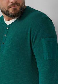 Zielona longsleeve koszula henley o teksturowanym wykończeniu, z okrągłym dekoltem, trzema guzikami i kieszonką naszywaną na lewym rękawie.