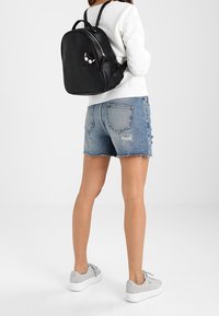 Czarny plecak z ekologicznej skóry o okrągłym kształcie, z przednią kieszenią na zamek i ozdobą w postaci zabawnych oczu. Noszony na białym swetrze i znoszonych jeansowych szortach.