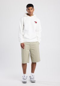 Sudadera blanca con bolsillo frontal y detalles gráficos rojos, combinada con pantalones cortos verde claro y zapatillas blancas con acentos grises.