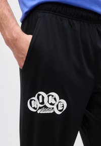 Αριστερό χέρι στην τσέπη μαύρου αθλητικού παντελονιού με λευκό λογότυπο «Nike Athletics», συνδυασμένο με μπλε πουκάμισο.