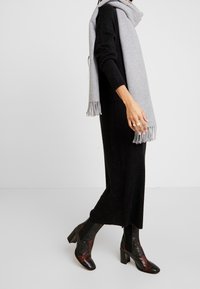 Femme portant une robe noire à manches longues, une écharpe grise à franges, des bagues aux doigts et des bottines à talons blocs noirs avec motifs, debout sur un sol blanc.