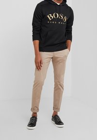 Person som bär en svart Hugo Boss hoodie, beige uppvikta byxor och svarta sneakers med vita sulor, stående mot en enkel grå bakgrund.