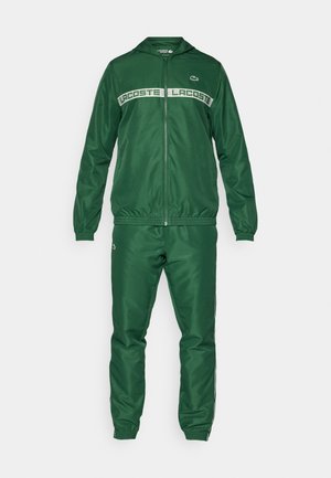 Tuta sportivo verde realizzato in materiale liscio e leggero. Presenta una giacca con zip e cappuccio, polsini elasticizzati e una fascia con logo sul petto.