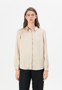 Beige ilgomis rankovėmis bluzonė, pagaminta iš satino audinio, su mygtukų užsegimu priekyje ir minkštais rankogaliais. Lygus paviršius su klasikinio apykaklės dizainu.