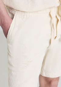 Primo piano di una mano di una persona nella tasca di pantaloni corti testurizzati di un bianco sporco con vita regolabile e una maglietta abbinata.