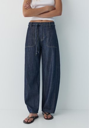 Kobieta w ciemnych dżinsach z wysokim stanem i szerokimi nogawkami, z przednimi kieszeniami, plecionym paskiem, białym bezrękawnikiem i czarnymi sandałami.