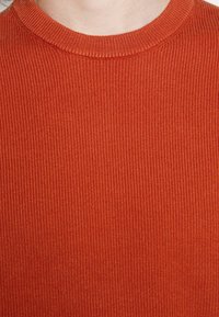 Gros plan d'une personne portant un pull à col rond côtelé de couleur orange brûlé, montrant la texture et le motif de tricot.