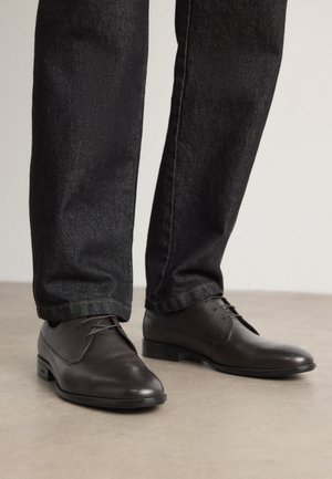 Dunkelbraune Lederschuhe mit glatter Textur und runder Zehenform, kombiniert mit schwarzen Denim-Jeans und einem klaren, minimalistischen Design.