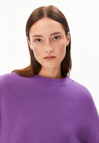 Junge Frau mit glatten braunen Haaren und Sommersprossen, die einen lila strukturierten Pullover trägt und direkt in die Kamera schaut, vor einem weißen Hintergrund.