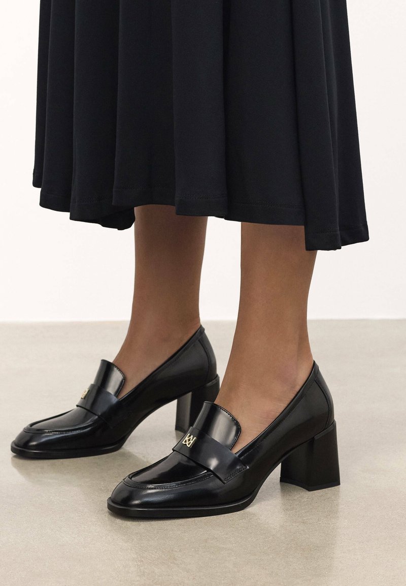 Mocassins en cuir verni noir avec un bout carré, un talon plat et un accent décoratif en matériel de teinte dorée. Texture lisse, design élégant.