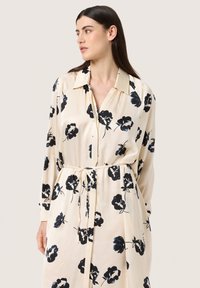 Rochie bluză de culoare crem cu imprimeu floral întunecat, mâneci lungi și talie legată. Dispune de un guler clasic și închidere cu butoane.