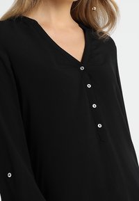Kvinde iført en sort langærmet bluse med V-udskæring og fire knapper langs fronten, samt en knapdetalje på ærmet.