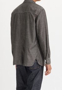 Grijze button-up shirt met lange mouwen, gemaakt van textuurstof. Heeft een gebogen zoom, een detail op de rugpas en knopenmanchetten. Draagt met donkere jeans.