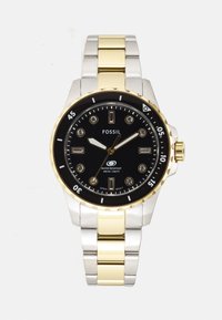 Fossil ur med sort urskive, bånd i sølv- og guldfarvet rustfrit stål, guld bezel og krystalmærker. Vandresistent op til 100m.