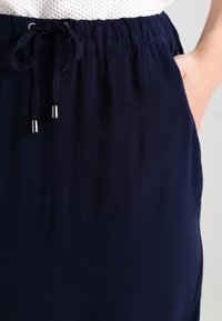 Tyrkysová sukně s elastickým pasem, šňůrkovým uzávěrem se stříbrnými koncovkami a bočními kapsami. Hladká textilie, ležérní design.
