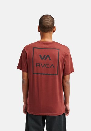 Mężczyzna o krótkich włosach, ubrany w rdzawy t-shirt z czarnym logo "VA RVCA" w kwadracie na plecach, stojący na jednolitym jasnym tle.