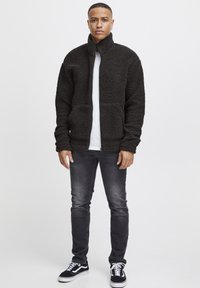 Giacca in pile nera e testurizzata con colletto alto e due tasche anteriori, abbinata a una camicia bianca, jeans grigi e sneakers nere.