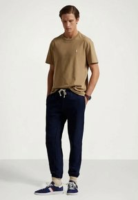 Camiseta de algodón caqui con cuello redondo, pantalones jogger azul marino con cordón y zapatillas azules con detalles blancos. Ajuste y diseño simples y casuales.