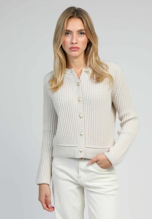 Jeune femme aux cheveux blonds portant un cardigan tricoté crème à boutons et un pantalon blanc, se tenant devant un fond clair et uni.