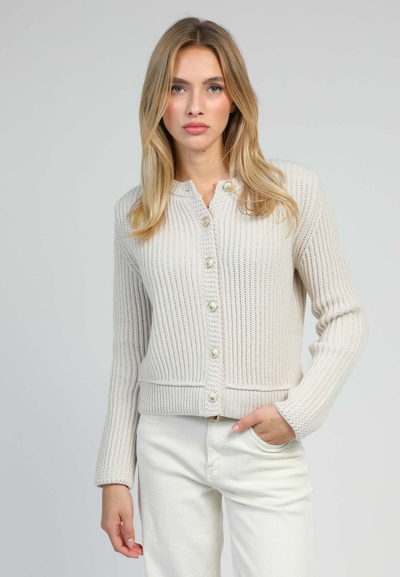 Jeune femme aux cheveux blonds portant un cardigan tricoté crème à boutons et un pantalon blanc, se tenant devant un fond clair et uni.