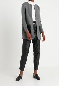 Nainen, jolla on päällään valkoinen poolopaita, harmaa auki oleva neuletakki mustilla taskuilla, mustat vyöllä varustetut nahkahousut ja mustat korokepohjaiset mokkasiinit.