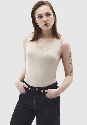 Giovane donna con capelli castani lisci che indossa un top beige senza maniche e jeans neri a vita alta, con le mani nelle tasche, guarda di lato.