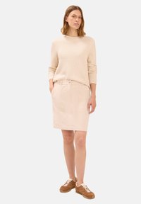 Žena stojící v béžovém texturovaném svetru, odpovídající sukni do délky ke kolenům a hnědých teniskách, s rukama v kapsách sukni.