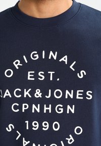 Námořnická modrá mikina vyrobená z bavlny s žebrovaným výstřihem. Obsahuje bílý text: "ORIGINALS EST. JACK & JONES CPNHGN 1990" v různých velikostech.