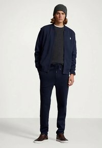 Jaunas vyras, apsirengęs tamsiai mėlynos spalvos treniruotėmis, tamsiai pilku megztiniu, pilku skrybėle ir rudos spalvos sportbačiais, stovi prieš paprastą baltą foną.